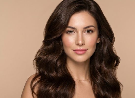 Retrato de una mujer con cabello castaño ondulado, piel radiante y fondo beige, representando belleza natural.